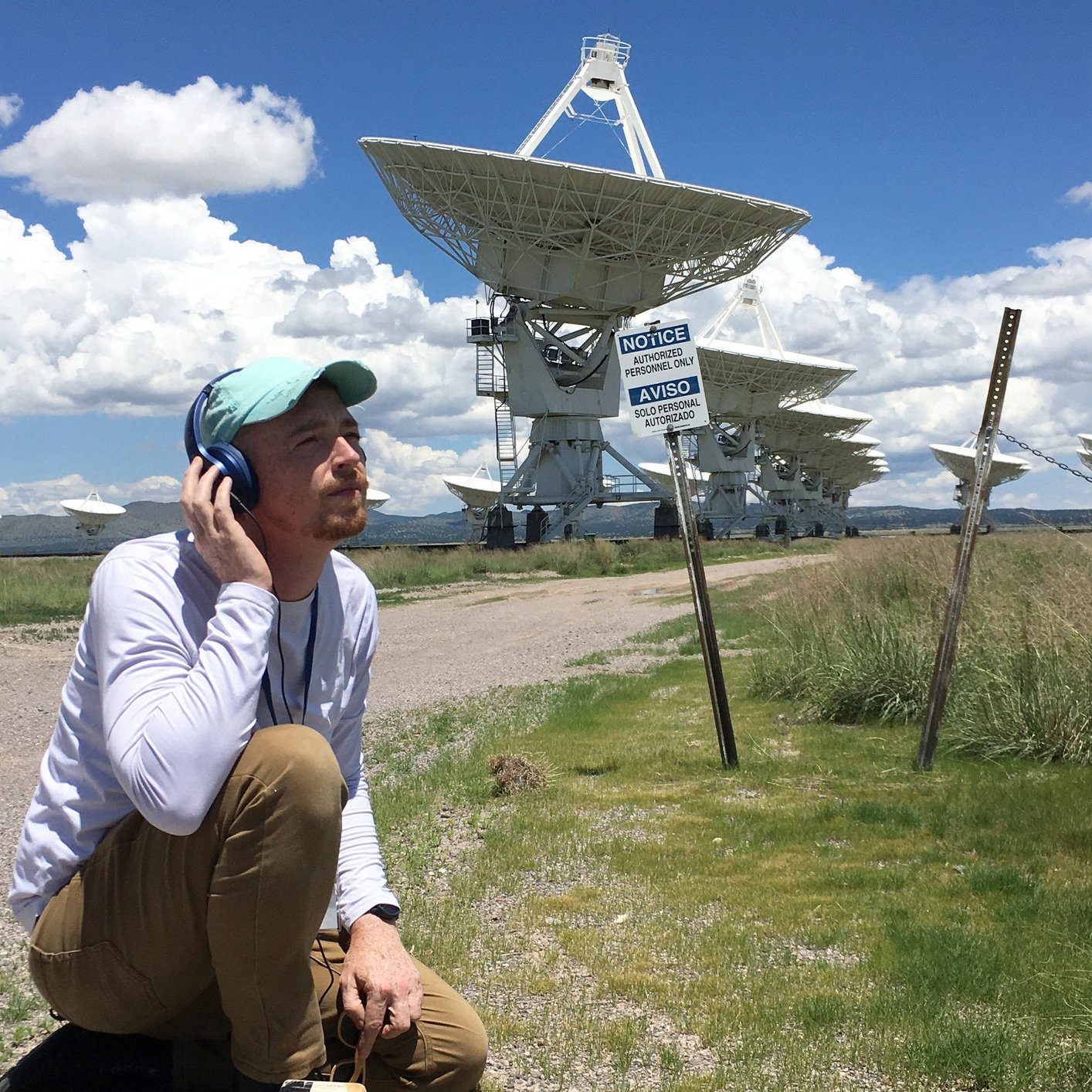 Photo of a man crouching in front of a series of massive radio dish antenna in the desert while holding a headphone to his ear in tribute to the movie poster for CONTACT